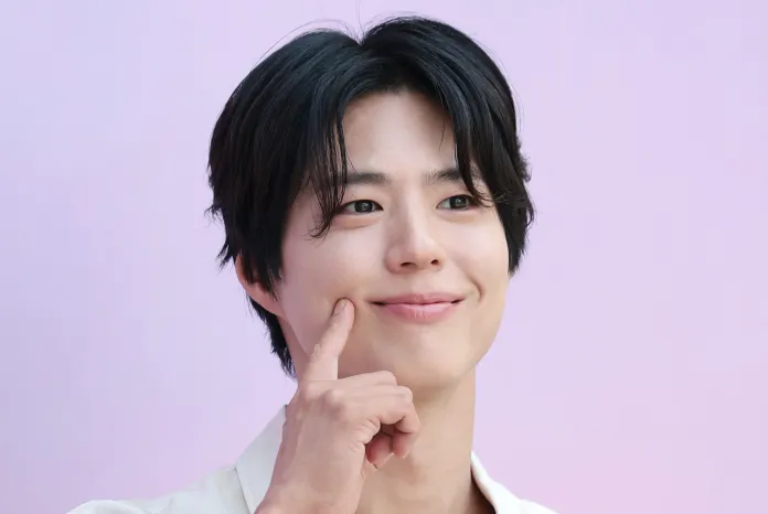 Park Bogum with black hair smiling and touching his cheek, set against a pink background.