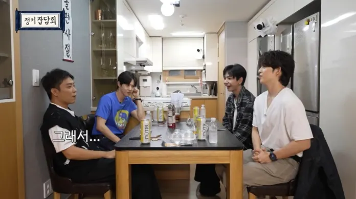 S.Coups sitting around a kitchen table, engaged in conversation. Three of them are smiling, and there are various drinks on the table, including cans and bottles.