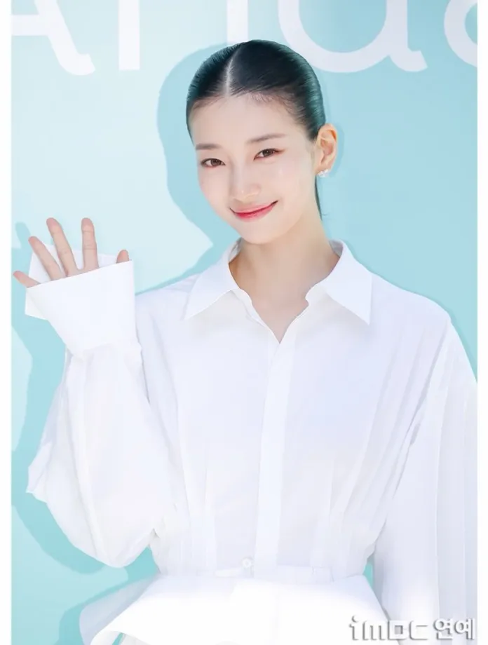 Suzy in a white oversized shirt smiling and waving, against a soft blue background.