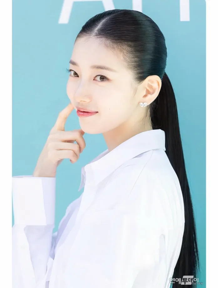 Suzy in a white shirt poses with a playful expression, touching her chin and smiling, against a light blue background.