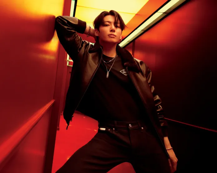 A stylish individual poses dramatically in a narrow red-lit hallway, wearing a black leather jacket and black pants, with accessories including chains and earrings.