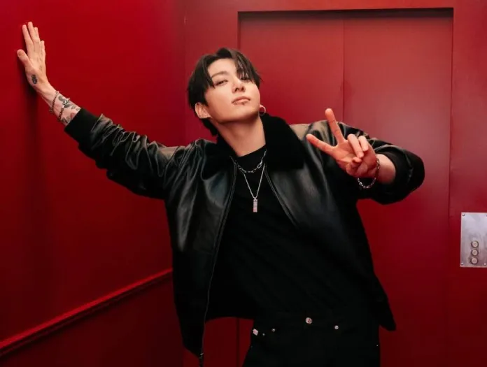 Jungkook  in a black leather jacket poses confidently against a red wall, making a peace sign with his right hand.