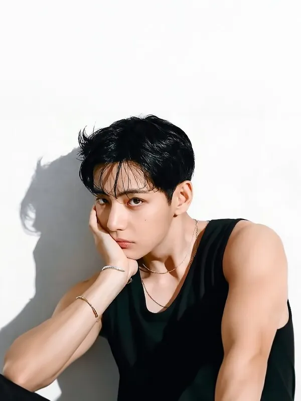 BTS V with short dark hair wearing a sleeveless black shirt, resting his head on his hand with a thoughtful expression. The background is bright and minimalistic.