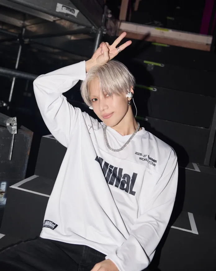 Taemin with light silver hair wearing a white long-sleeve shirt with text and a silver chain, sitting on stairs and making a peace sign with one hand.