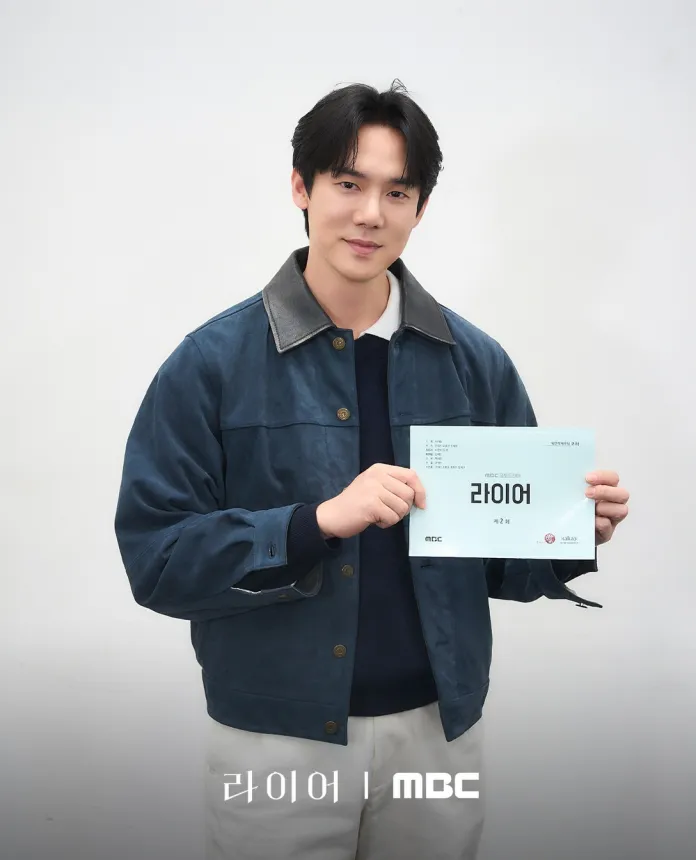 Yoo Yeon-seok dressed in a blue jacket and sweater is smiling while holding a script for a project titled '라이어' against a plain white background.