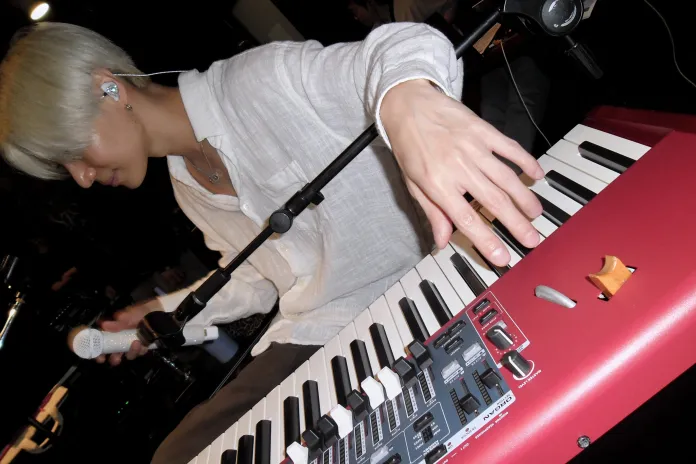 Taemin with silver hair is playing a red keyboard while holding a microphone, in a performance setting.