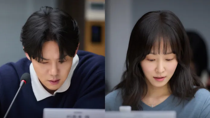 A focused man and woman in a meeting, both looking down at documents in front of them. The man wears a dark sweater and the woman a light blue top.