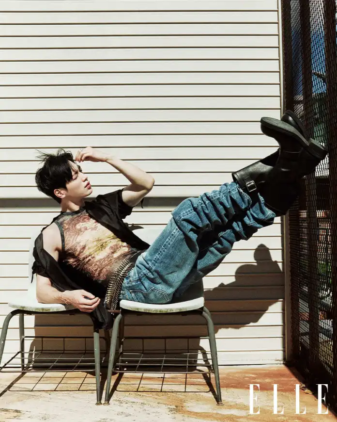 Seo Kang-joon reclining on a chair against a backdrop of wooden slats, wearing a graphic tank top and loose denim jeans, with one leg extended and styled black boots, in a relaxed pose.