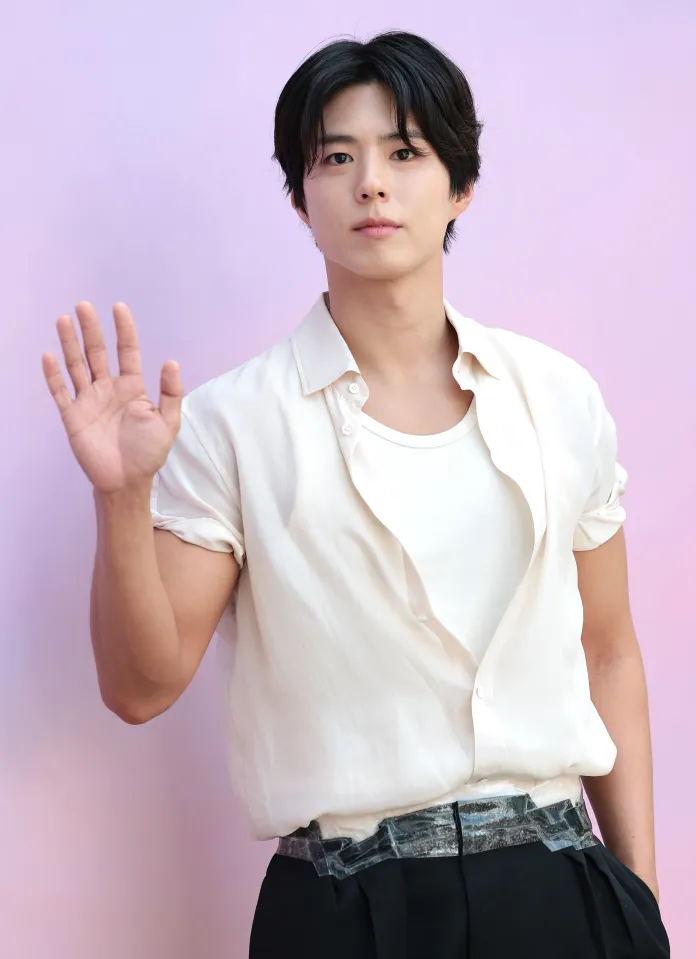 Park Bo-gum with dark hair wearing a white shirt and black trousers is standing against a pastel purple background, raising his right hand in a wave.