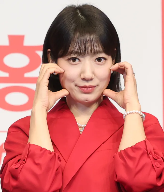 Park Shin-hye in a red outfit is making a playful gesture with her hands on her cheeks, smiling at the camera.