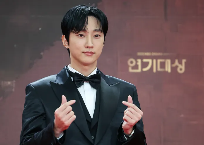 Jinyoung in a black tuxedo and bow tie poses confidently, making heart gestures with his hands at a red carpet event.