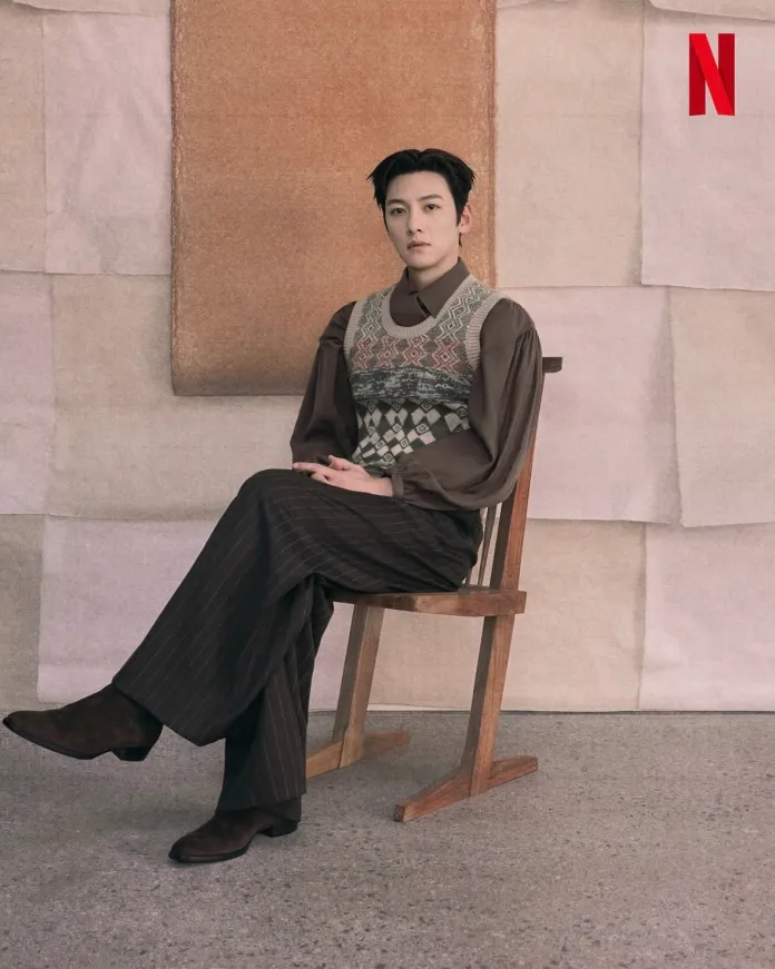 Ji chang wook seated on a wooden chair against a textured background made of various colored paper. They are wearing a patterned vest over a brown shirt and striped pants, paired with dark footwear.