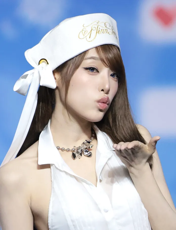 YENA poses with a playful expression, blowing a kiss while wearing a white outfit and unique headwear.