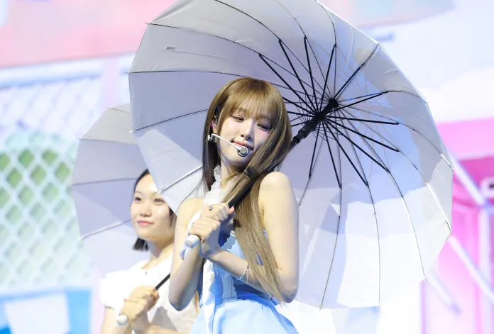 YENA in a light blue outfit holding a white umbrella on stage, with another person smiling in the background.
