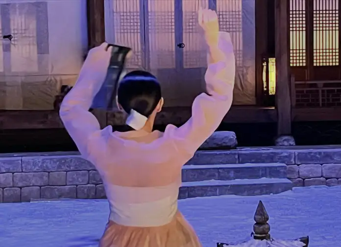 Son Ye-jin in traditional attire raises an object above their head, with a serene outdoor setting illuminated by soft light and a snow-covered ground.