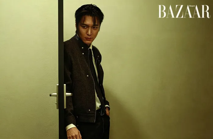 Lee Min-ho stands by a partially open door, dressed in a stylish dark jacket and tie, against a muted green wall.