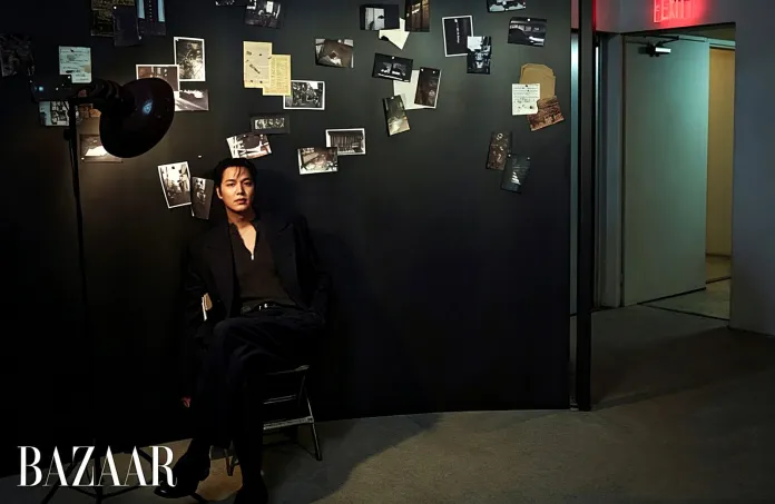 Lee Min-ho in a black outfit sitting on a chair against a dark wall decorated with various photographs and papers, with a spotlight illuminating him.