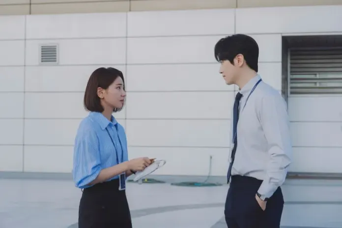 A young woman in a blue shirt holds a clipboard while talking to a man in a white dress shirt and tie in a modern outdoor office setting.