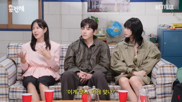 Son Ye-jin, Ji chang wook and NANA sitting on a plaid couch engaged in conversation, with one person gesturing expressively. They are wearing casual, stylish clothing and there are colorful cups in front of them.