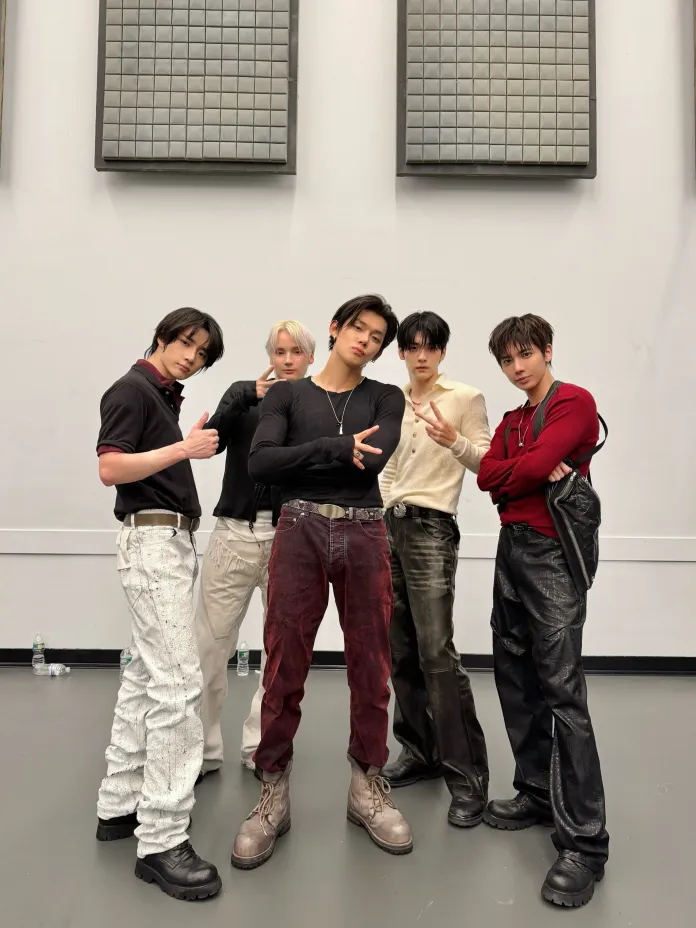 TXT members posing together indoors, dressed in trendy outfits with varying styles. They are standing confidently in a stylish formation, showcasing different hairstyles and expressions.