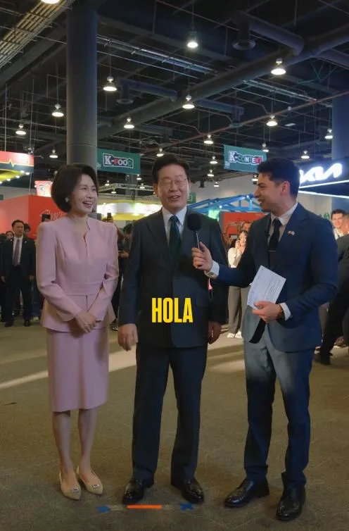 Korean President and his wife stand in a brightly lit exhibition space, with a colorful backdrop. Christian Burgos is holding a microphone while interviewing another, who is smiling. The third person is smiling and looking at the other two.