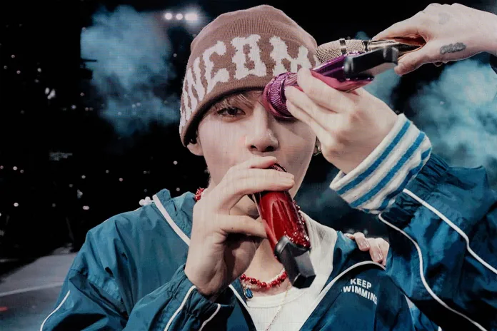 BTS V on stage holding a microphone while someone off-camera assists with another microphone. The performer is wearing a brown beanie and a blue jacket.