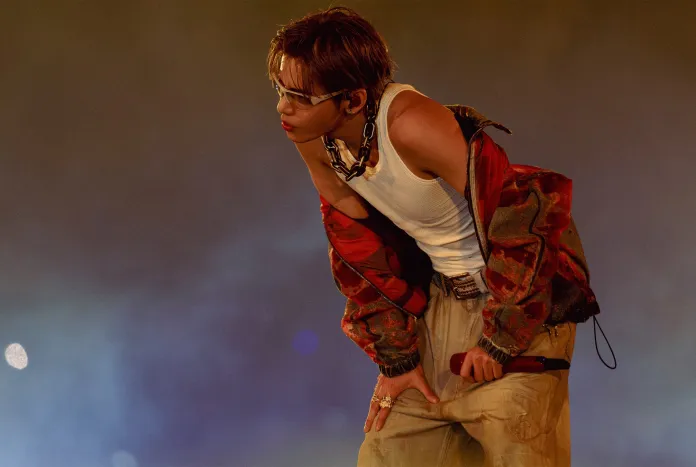 BTS V wearing a white tank top and a colorful jacket, holding a microphone and posing with an expressive expression.