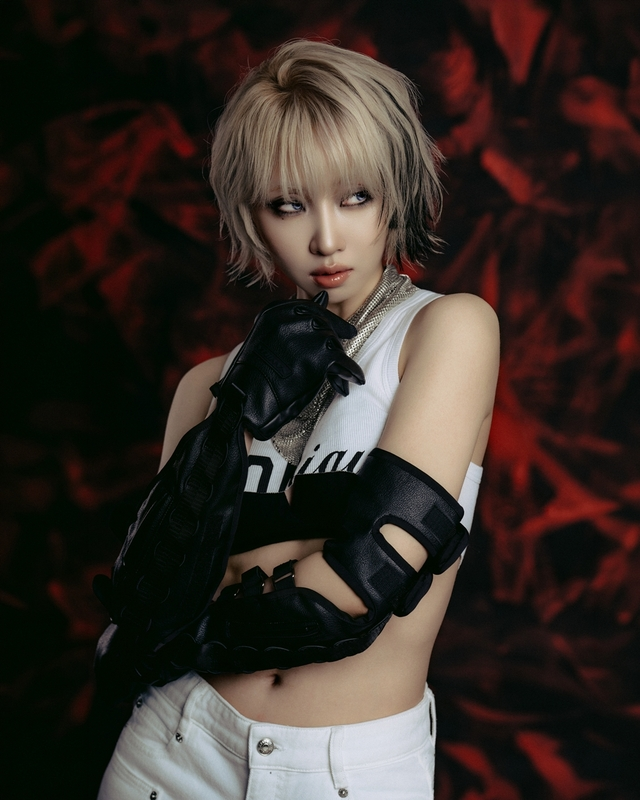 A stylish individual with short, layered blonde hair wearing a sleeveless white top and black gloves, posing against a dark, abstract red background.