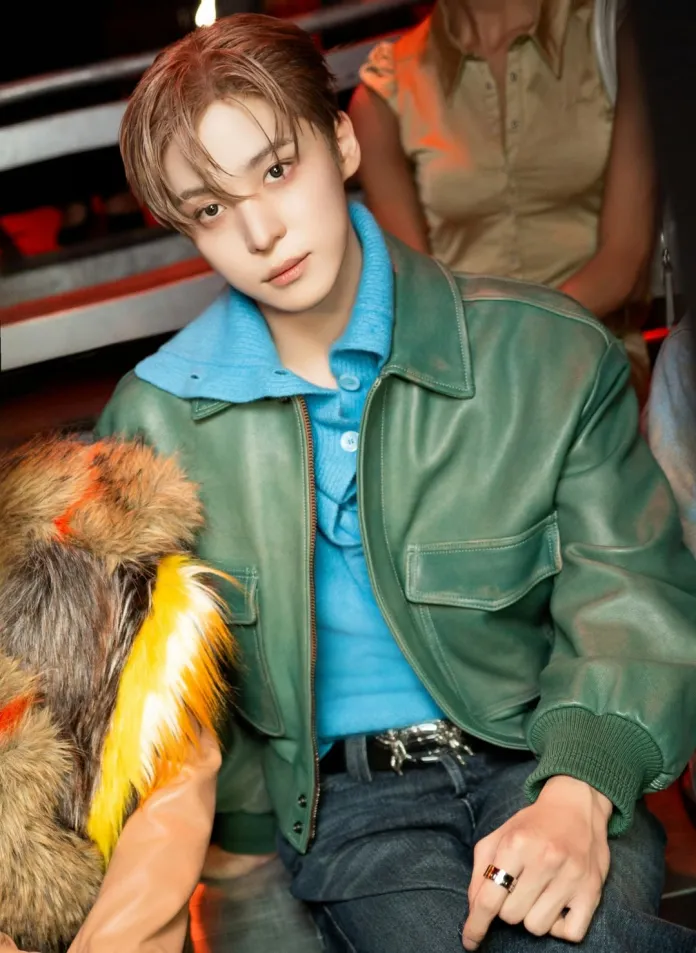 Yunho with styled hair wearing a green leather jacket over a blue sweater, seated in a relaxed pose with one hand on their knee, and colorful fur on the side.