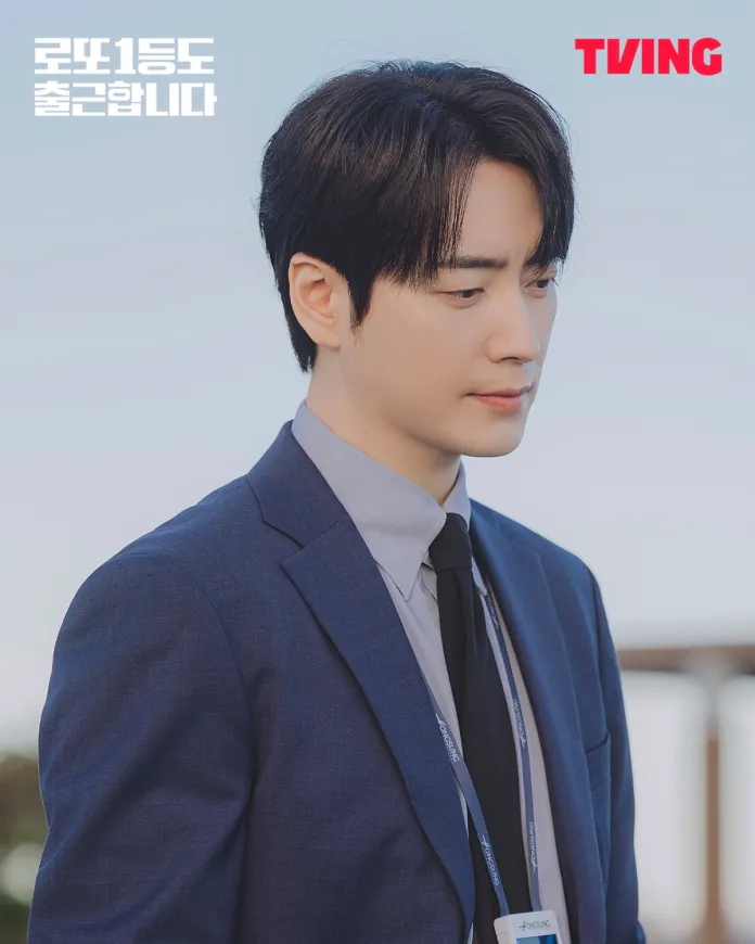 Lee Joon-hyuk wearing a blue suit and tie, looking down thoughtfully. He has dark hair and is standing outdoors with a light sky in the background. A TVING logo is visible in the top right corner.