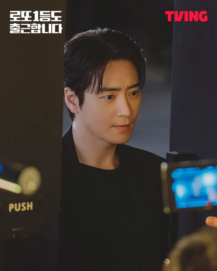A close-up shot of Lee Joon-hyuk with dark hair, wearing a black shirt and jacket, looking intently with a serious expression. A camera is visible in the foreground capturing the moment.