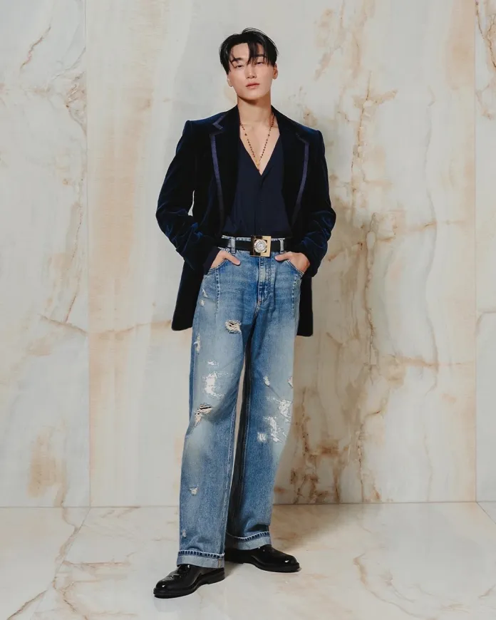 San poses confidently against a marble backdrop, wearing a dark velvet blazer over a deep v-neck shirt and distressed jeans, complemented by stylish black shoes and accessories.