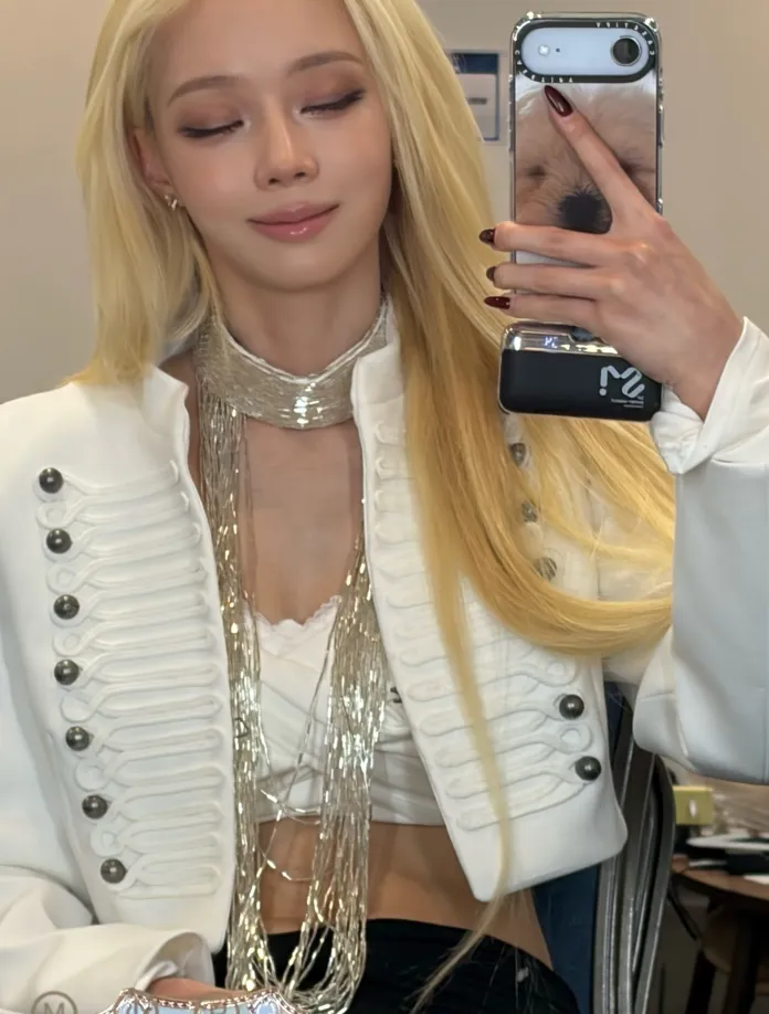 Winter with long blonde hair wearing a stylish white jacket and a shimmering necklace, taking a selfie with a phone.