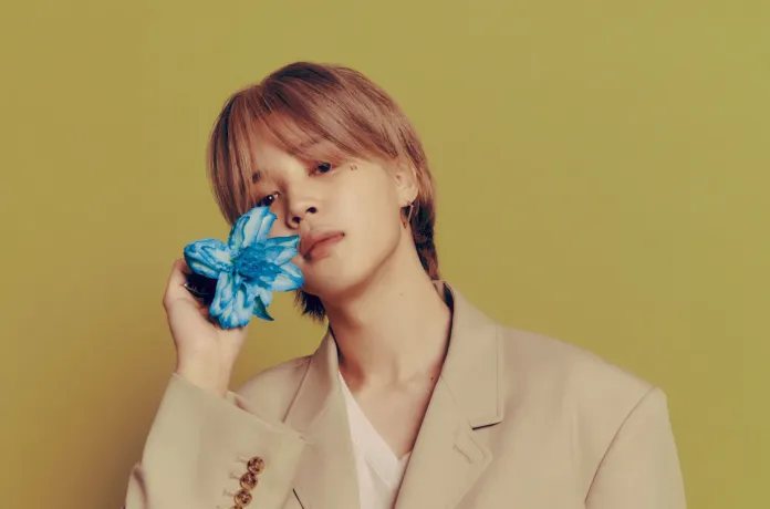 Jimin with light brown hair wearing a beige oversized blazer, holding a blue flower close to their face against a green background.