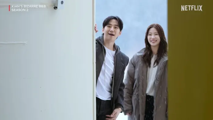 Lee Junho and Kazuha standing at an open door, covered in light snow, with a winter landscape visible in the background. The scene captures a cheerful moment from a show.