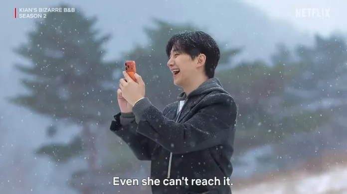 Lee Junho smiling and holding a phone while taking a photo in a snowy landscape, with trees softly blurred in the background.
