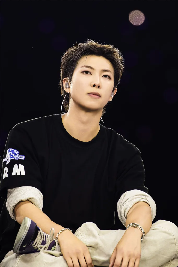 RM with short hair sitting on stage, wearing a black shirt and light-colored pants, with a serious expression and microphone in his ear.