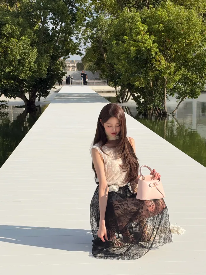Wonyoung with long hair is sitting on a white wooden walkway surrounded by greenery. She is wearing a light top and a black lacy skirt, holding a small pink handbag while looking down.