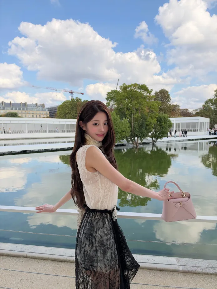 Wonyoung standing by a reflective body of water, wearing a light-colored top and a black lace skirt, holding a small pink handbag, with trees and a cloudy sky in the background.