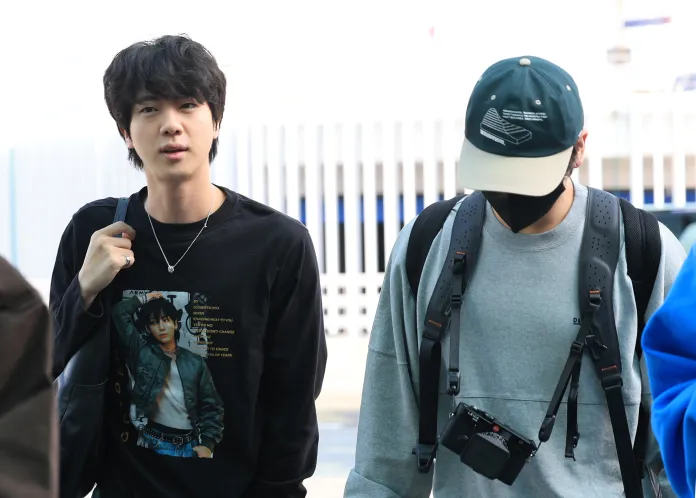 Two individuals walking together; one in a black long-sleeve shirt with a graphic print and the other wearing a gray sweatshirt and a cap. The second person is partially obscured by a mask and has a camera hanging from their shoulder.