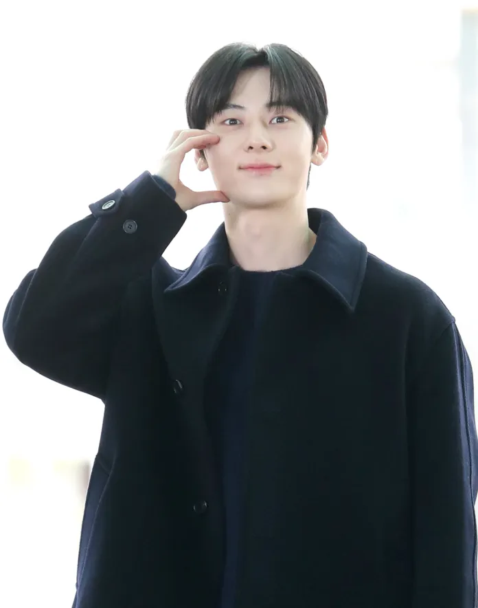 Hwang Min-hyun smiling and making a playful gesture with his hand while wearing a dark coat.