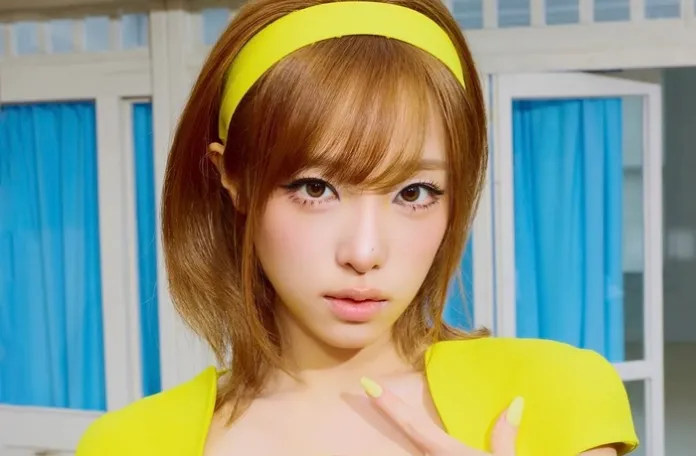 YENA with short brown hair wearing a yellow headband and a yellow outfit, posing in front of a light background with blue curtains.