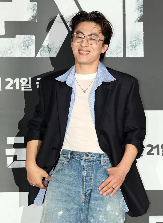 A smiling Koo Kyo-hwan wearing glasses, a black blazer over a white shirt with a blue collar, and distressed denim jeans, posing in front of a promotional backdrop.