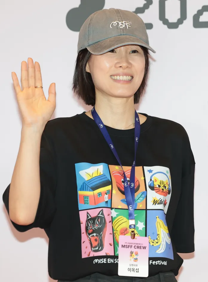 Lee Ok-seop  with short black hair, wearing a gray cap and a colorful graphic t-shirt, waves while standing in front of a banner for a film festival.
