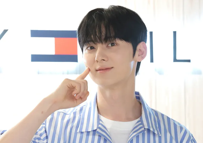 Hwang Min-hyun with black hair, wearing a striped blue and white shirt, smiling and resting a finger on their cheek against a softly blurred background.