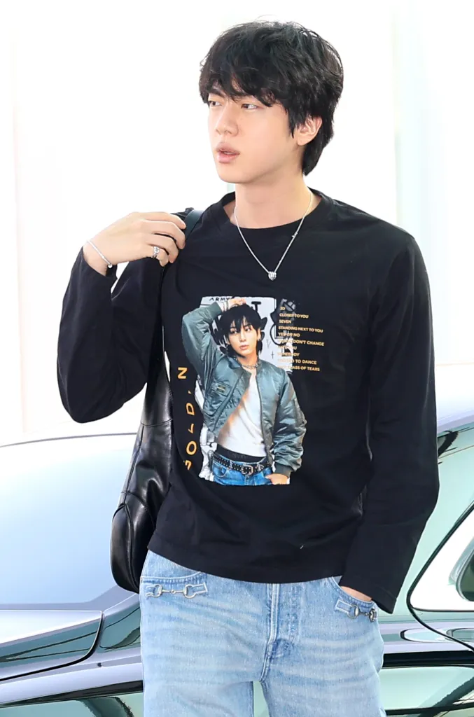 Jin with dark hair wearing a black long-sleeve shirt featuring a photo print and light blue jeans, holding a shoulder bag while looking slightly away.