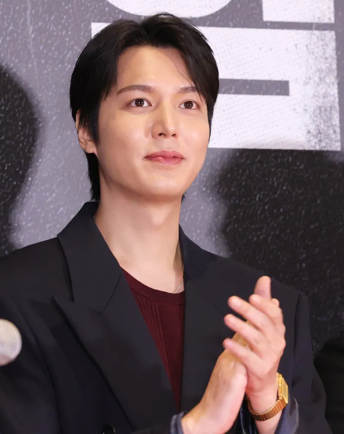 Lee Min-ho with dark hair and a thoughtful expression, wearing a stylish blazer and a reddish sweater, is clapping at a public event.