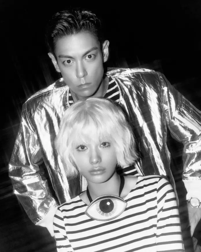 A black-and-white image of T.O.P and Nana, T.O.P with slicked-back dark hair wearing a shiny silver jacket, and Nana with short, wavy blonde hair in a striped shirt and an eye-shaped necklace.