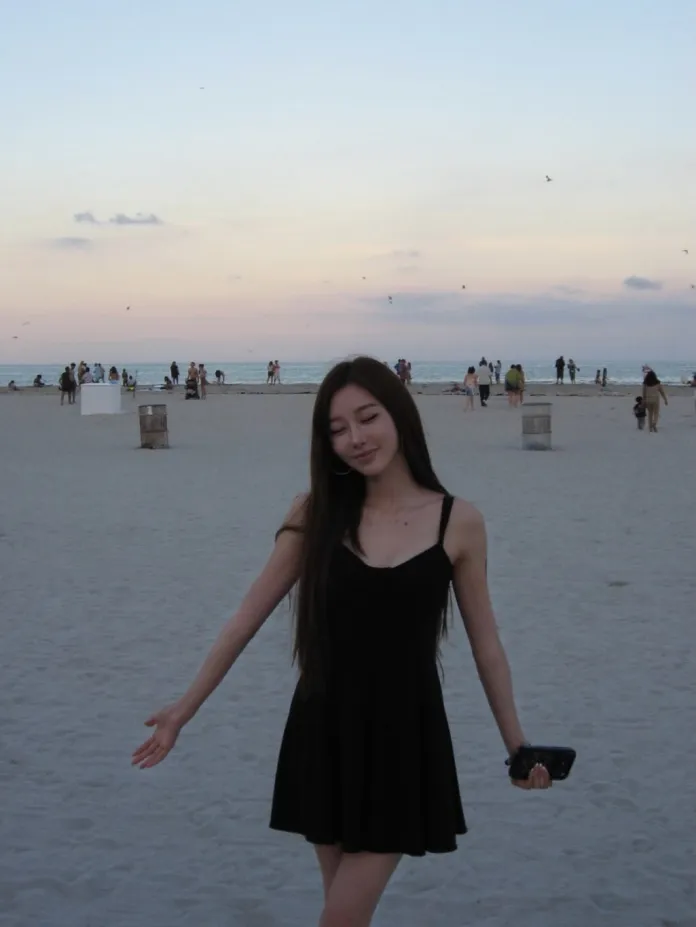 Giselle  in a black dress standing on a sandy beach at sunset, with the ocean and people in the background.