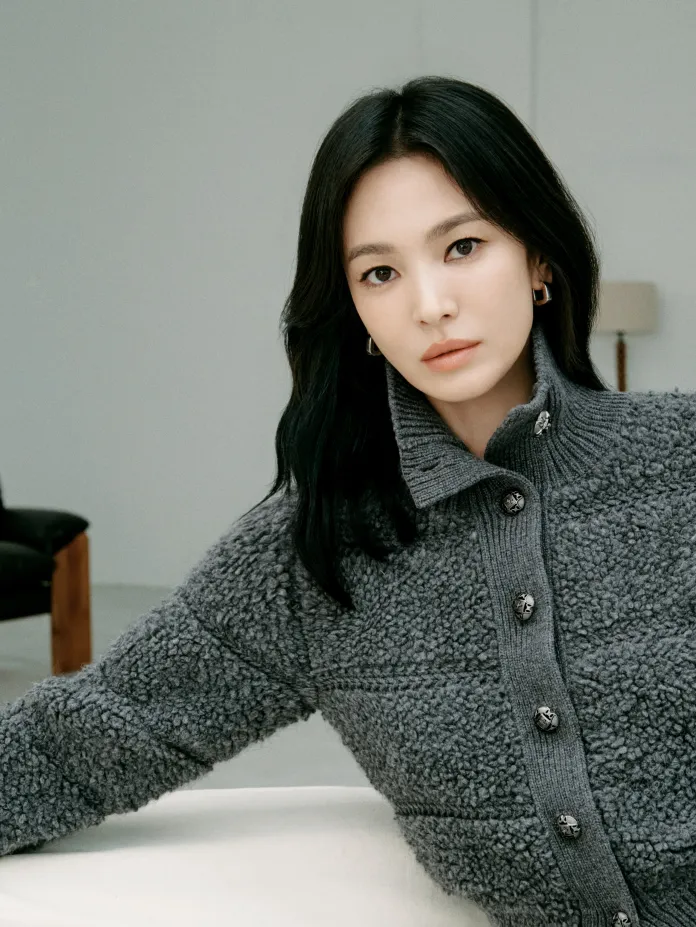 Song Hye-kyo with long black hair, wearing a cozy gray cardigan, is seated casually and looking directly at the camera with a neutral expression in a minimalist indoor setting.